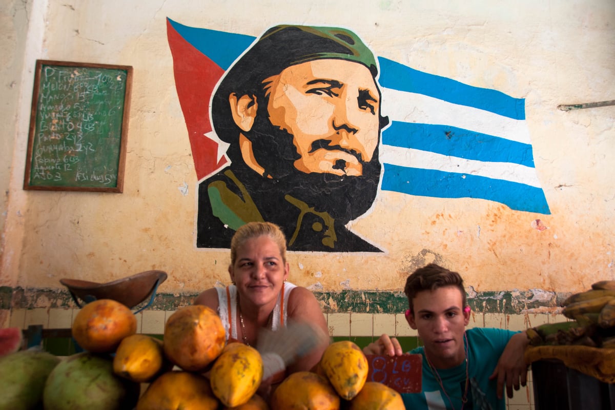 ritratto di una donna e di un ragazzo cubani, fronte a loro della frutta, sullo sfondo un murales con il volto di Fidel Castro e la bandiera cubana