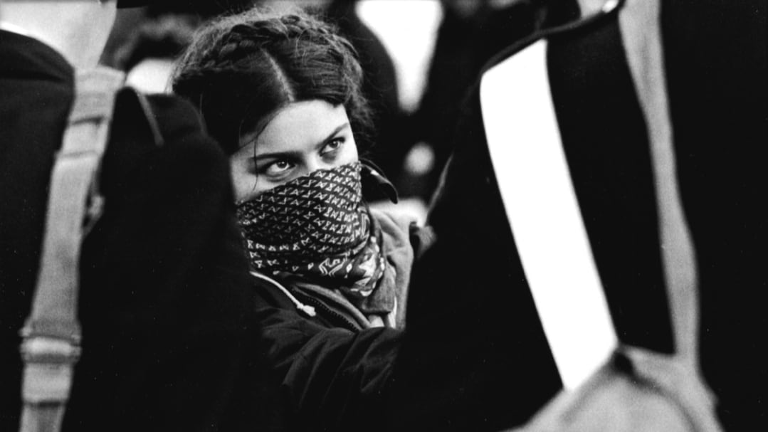 Una ragazza con il volto coperto da una sciarpa guarda intensamente dei carabinieri di fronte a lei. - Foto di Tano D'amico