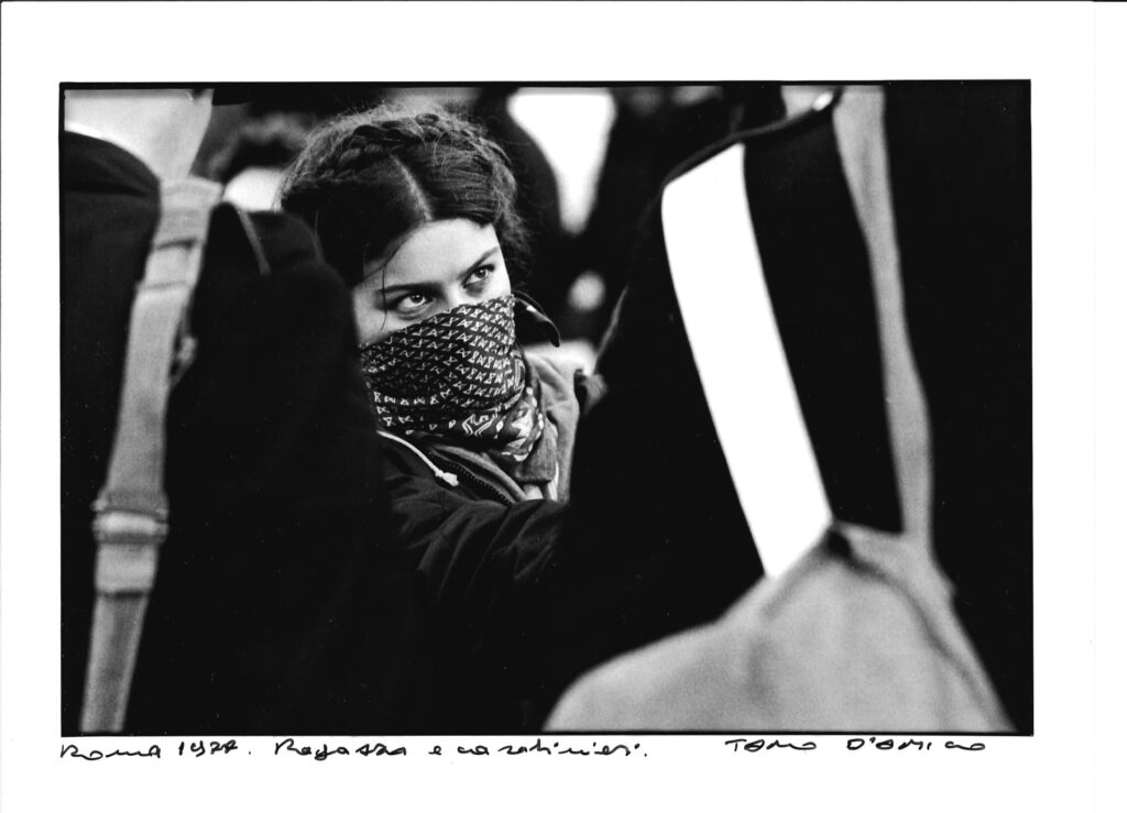 Una ragazza con il volto coperto da una sciarpa guarda intensamente dei carabinieri di fronte a lei. - Foto di Tano D'amico